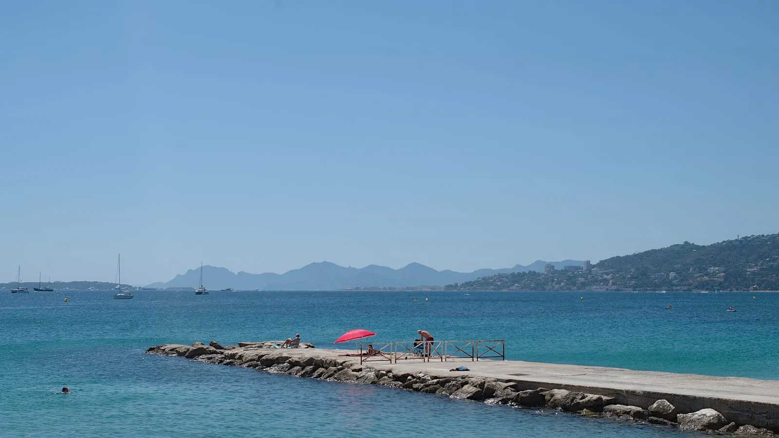 La baie de Juan-les-Pins : eau turquoise, ponton de baignade, voiliers au mouillage et massif de l'Estérel à l'horizon