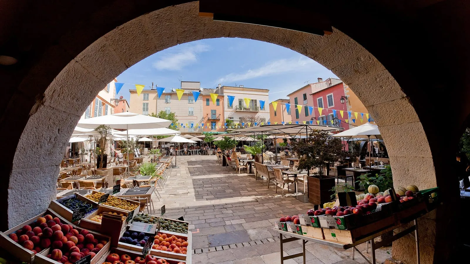 La place des Arcades au cœur du village de Valbonne, vue sous une arche en pierre, avec ses étals de marché provençal et ses terrasses de café